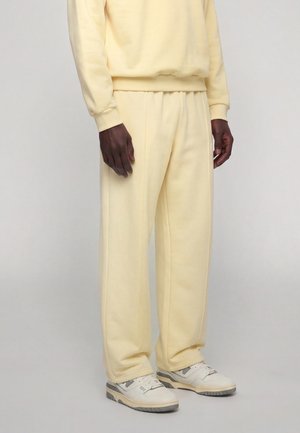Homme portant un pantalon de survêtement couleur crème et un sweat-shirt avec des baskets blanches et grises, debout devant un fond clair uni.