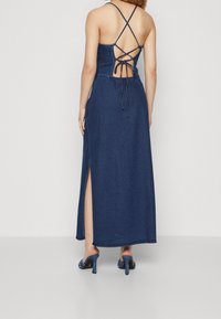 Robe maxi en denim avec un corsage ajusté, des bretelles croisées à l'arrière et des fentes latérales ; couleur bleu foncé ; texture lisse ; associée à des sandales à talons bleues.