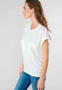 Top corto bianco a maniche corte con due tasche frontali e dettagli di cucitura sottili sulle maniche. Abbinato a jeans in denim blu. Texture del tessuto liscia.