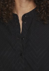 Haut noir boutonné avec un motif en chevrons, tissu texturé et un décolleté peu profond avec deux boutons noirs visibles.