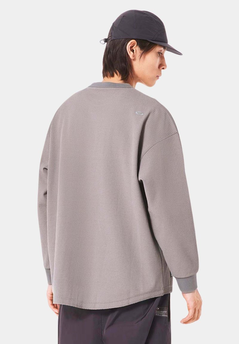 Gray, ribbed long-sleeve shirt with a relaxed fit; features grey cuffs and a small logo on the back shoulder. Worn with a contrasting cap.