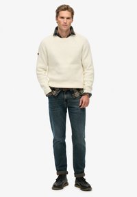 Junger Mann steht mit den Händen in den Taschen, trägt einen cremefarbenen Pullover über einem karierten Hemd, blaue Jeans, schwarze Stiefel und eine Armbanduhr.