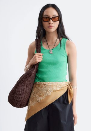 Femme portant un haut vert sans manches, une écharpe marron à motifs nouée à la taille, un pantalon noir, un sac marron texturé et des lunettes de soleil rectangulaires marron.