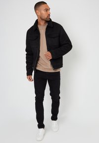 Sort ruskindjakke med et shearling-krave, to brystlommer, båret over en beige top, parret med sorte jeans og hvide sneakers.