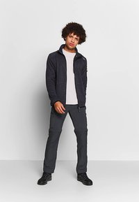 Ung mand med krøllet hår iført en mørk zip-up jakke, hvid t-shirt, mørke bukser og sorte sneakers, stående mod en simpel lys baggrund.