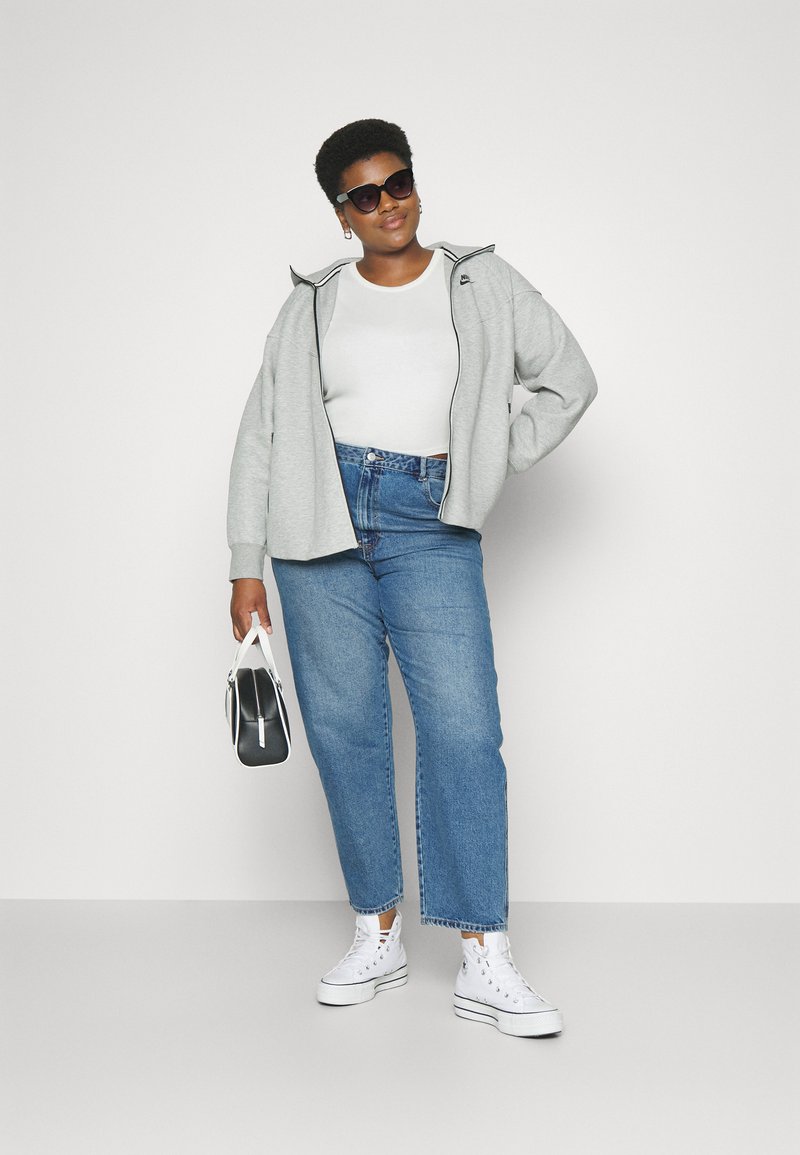 Haut de survêtement gris oversize avec fermeture éclair, top blanc à col rond, jeans bleus taille haute, baskets blanches, et sac à main noir avec accents blancs.