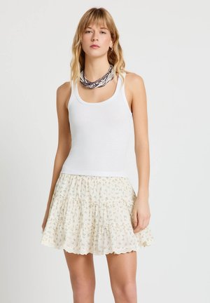 Donna che indossa una maglia bianca senza maniche, gonna crema a fiori con balze e collana a strati di perline, in piedi contro uno sfondo semplice.