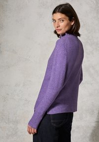 Lila, strukturiertes Strickoberteil mit hohem Kragen, gerippten Bündchen und seitlichen Schlitzen, kombiniert mit dunklen Jeans. Weiches, kuscheliges Material.