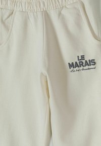 Pantalon de survêtement couleur crème avec taille élastique et poches latérales, avec le texte "LE MARAIS Le 3e Arrondissement" sur la cuisse droite.