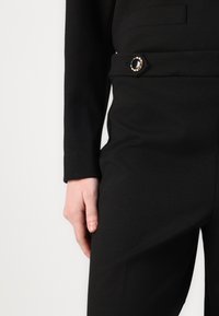 Zwarte aansluitende broeken van een gestructureerde stof, met een decoratieve zilveren knop en een gestileerde tailleband, subtiel afgewerkte zakken.