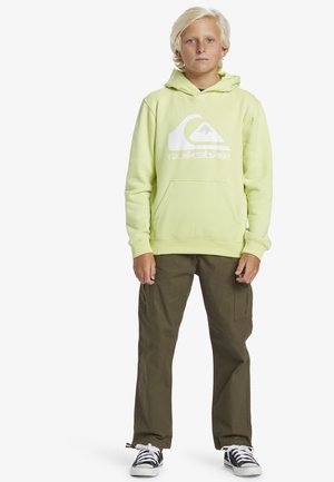 Felpa verde lime con logo bianco, tasca frontale; abbinata a pantaloni cargo oliva, con tasche laterali e vestibilità comoda; sneakers in bianco e nero.