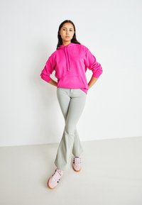 Felpa rosa con cappuccio e cordini, tasca frontale, abbinata a jeans svasati grigi chiari e sneakers rosa con accenti più scuri.