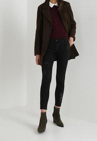 Svarta tajta jeans med slitna detaljer, parat med en vinröd tröja och olivgröna ankelstövlar. Lager på en mörkbrun kappa.