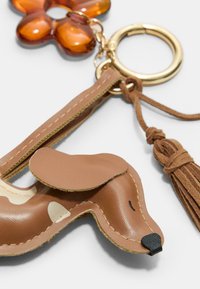 Brun læder hundeformet nøglering med en syet tekstur, der har en gylden nøglering og frynse-detalje. En ravfarvet blomst charm er fastgjort.