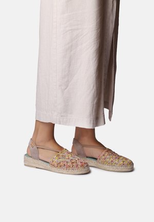 Nogi w beżowych spodniach o szerokich nogawkach oraz w wielokolorowych plecionych espadrylach z paskami wokół kostek na białym tle.