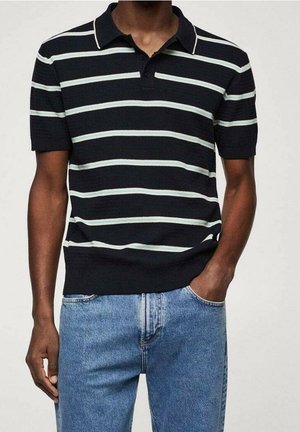 Homme portant un polo noir à rayures blanches horizontales et un jean bleu clair, une main dans la poche, sur fond uni.
