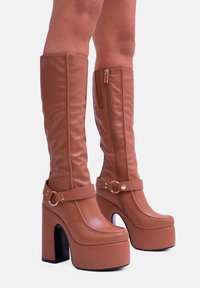 Stivali alti in finta pelle marrone, con piattaforma, chiusura laterale con zip, cinturino alla caviglia e punta tonda. Presentano un tacco alto chunky e una superficie liscia.