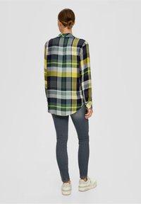 Langærmet skovmandskjorte i grønne, gule og marine farver; knapmanchetter; rund kant; båret med mørke jeans og hvide sneakers.