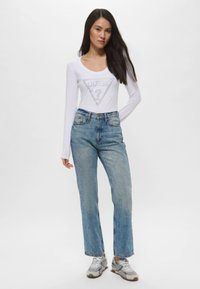 Maglia bianca a maniche lunghe con logo triangolare argento "GUESS?", abbinata a jeans azzurri a vita alta e sneakers grigie.