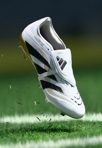 Baltie futbolsneakeri ar melnām un zeltītām svītrām, ar plūstošu dizainu, teksturētu virsu un izteiksmīgu jostu vidusdaļā.
