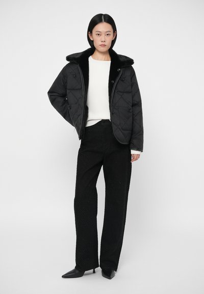 Μαύρη καπιτοναρισμένη ζακέτα με απομίμηση γούνας, συνδυασμένη με λευκό τοπ και μαύρα υφασμάτινα παντελόνια. Το μοντέλο στέκεται μπροστά σε απλό φόντο.