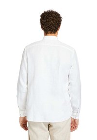 Camicia bianca in lino a maniche lunghe con yoke posteriore, polsini con bottone e orlo leggermente curvo, che mette in evidenza una texture leggera e traspirante.