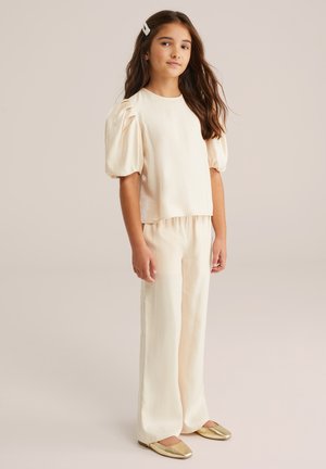 Mädchen mit langen braunen Haaren, das ein cremefarbenes Oberteil mit Puffärmeln, passende weite Hosen, goldene Ballerinas und eine Perlenhaarnadel trägt, steht vor einem beigen Hintergrund.