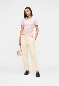 Γυναίκα που στέκεται φορώντας ροζ ριγέ μπλουζάκι, κρεμ παντελόνι φαρδιάς γραμμής, μαύρα μοκασίνια, κρατώντας μπεζ τσάντα χειρός μπροστά σε λευκό φόντο.