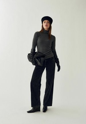Femme portant un béret noir, un col roulé gris, des gants noirs, un pantalon large noir et des bottes noires, tenant une veste en cuir noire pliée.
