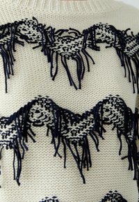Cremfarbener Strickpullover mit marineblauen dekorativen Fransen und Zickzackmuster. Die Textur ist weich, mit ausgeprägten gewebten Details und offenen Strickbereichen.