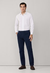 Camisa blanca de botones, pantalones azul marino, zapatos marrones. La camisa tiene un cuello puntiagudo; los pantalones son de pierna recta con bolsillos frontales y lazos para cinturón.