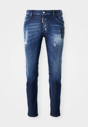 Jeans in denim blu scuro con vestibilità slim. Presentano dettagli strappati e una leggera scoloritura. Include una toppa a contrasto nell'area della tasca.