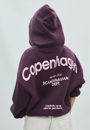 Person iført en lilla hættetrøje med hætten oppe og tekst "Copenhagen," "SCANDINAVIAN DEPT.," og "Creativity never asks for permission" på ryggen.