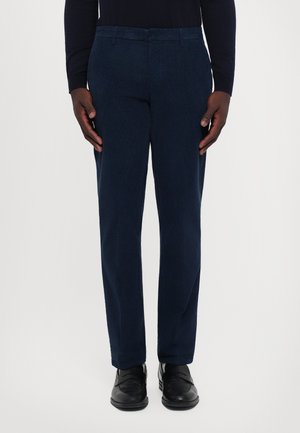 Mann trägt marineblaue gerade geschnittene Hose, schwarze Leder Slipper und ein dunkles langärmeliges Oberteil, steht vor einem weißen Hintergrund.