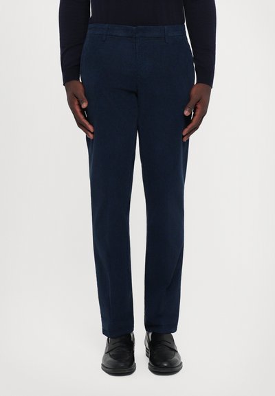 Homme portant un pantalon droit bleu marine, des mocassins en cuir noirs et un haut à manches longues foncé, se tenant contre un fond blanc.