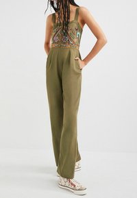 Olivgrön overall med justerbara band, broderad urringning med färgglada mönster, vida ben och sidofickor. Bärs med mönstrade sneakers.