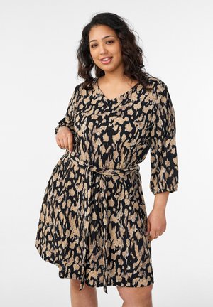 Žena s kučeravými vlasmi, oblečená v čiernych a béžových šatách so zvieracím vzorom, s tromištvrtinovými rukávmi a opaskom viazaným v páse, usmieva sa do kamery.