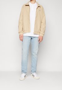 Beige leichte Jacke mit elastischen Bündchen, weißes Polohemd, hellblaue Jeans und weiße Sneakers mit schwarzen Streifen. Einfaches, lässiges Outfit.
