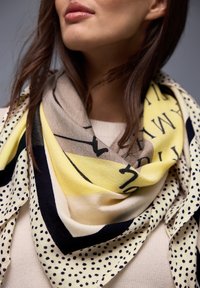 Femme portant un pull crème et une écharpe jaune, noire et beige à motifs de lettres et de pois autour du cou.