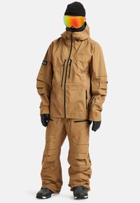 Brązowa wodoodporna kurtka i spodnie śniegowe, z regulowanymi mankietami, wieloma zapinanymi na zamek kieszeniami i teksturowanym panelem. Noszone z rękawiczkami i goglami.