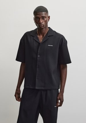 Ung mand iført en sort kortærmet skjorte med knapper og matchende shorts står mod en ensartet lys grå baggrund.