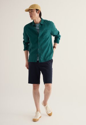 Uomo che indossa un berretto beige, una camicia verde scuro abbottonata, pantaloncini blu scuro e scarpe da ginnastica bianche, in piedi davanti a uno sfondo chiaro uniforme.