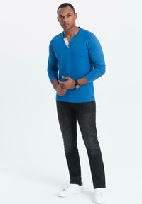 Blaues Langarm-Henley-Shirt mit drei Knöpfen, kombiniert mit dunklen Jeans und weißen Sneakern. Das Shirt hat eine glatte Textur und einen lässigen Schnitt.
