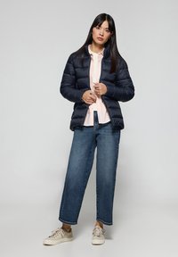 Giubbotto imbottito blu con colletto alto, indossato sopra una camicia rosa chiaro e jeans a gamba larga blu. Sneakers beige completano l'outfit.