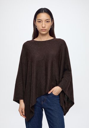 Young woman with long dark hair wears loose brown poncho and blue jeans, standing with one hand in pocket against plain background.