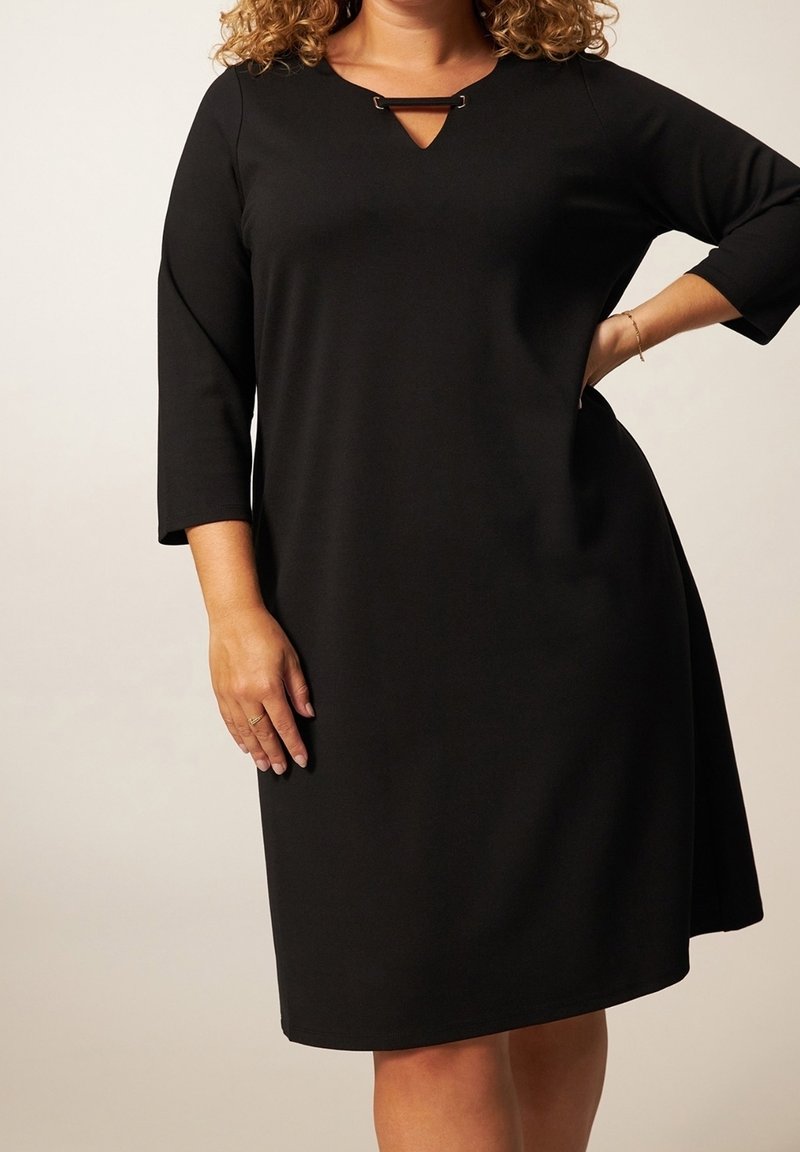 Femme portant une robe noire longueur genou avec des manches trois-quarts et une petite découpe triangulaire au niveau du décolleté avec un détail en barre métallique.