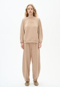 Beige sweatshorts och lösa byxor i mjukt tyg, med en präglad logotyp på bröstet och ribbade muddar. Modellen står rakt.