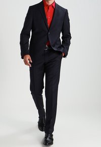 Dunkler maßgeschneiderter Anzug mit tailliertem Sakko und gerade geschnittenen Hosen, kombiniert mit einem roten Hemd mit Knopfleiste und schwarzen Schuhen, vor einem neutralen Hintergrund stehend.