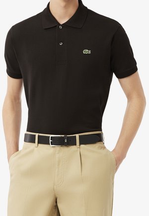 Mann trägt schwarzes Poloshirt mit kurzen Ärmeln und grünem Krokodil-Logo sowie beige Hose mit schwarzem Gürtel, Hände in den Taschen.