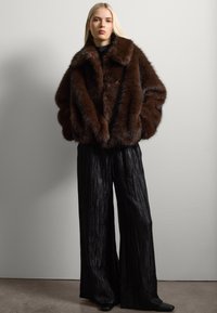 Chaqueta de piel sintética marrón con un amplio cuello, combinada con pantalones de pierna ancha plisados negros. Texturas suaves, gran calidez y un rico contraste de color.